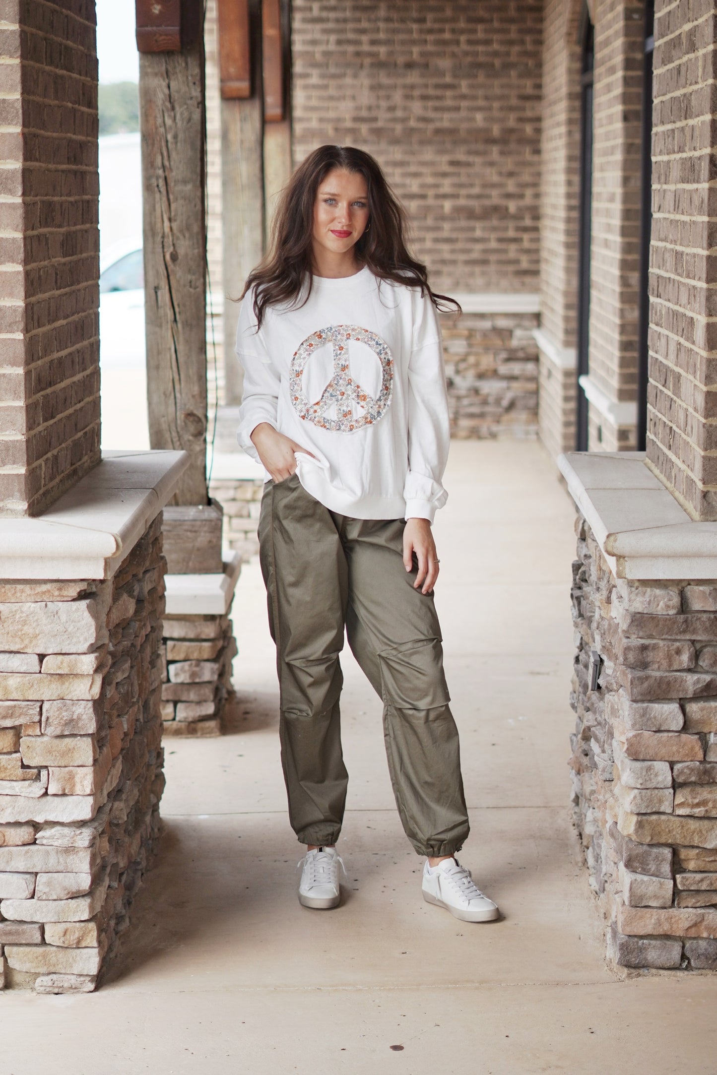 Polly Parachute Jogger Pants Elastic Waistband w/ Zipper Fly and Button Closure Full Length Legs Cinched at the Ankle Pockets Dark Olive Color. Brunette model, white sweatshirt with peace sign, sneakers. Stone and brick background.