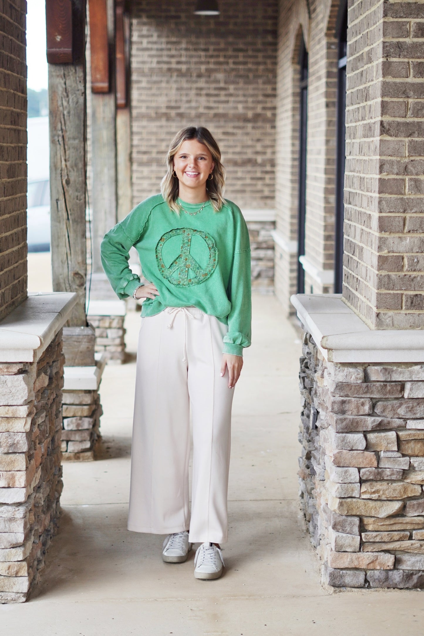 Remi Raw Hem Peace Sign Graphic Pullover Crew Neckline Long Sleeve Peace Sign On Front Of Top Colors: Green, Off White Relaxed Fit Full Length. Blonde model, green version of shirt, cream wide leg sweats, sneakers, stone and brick background.