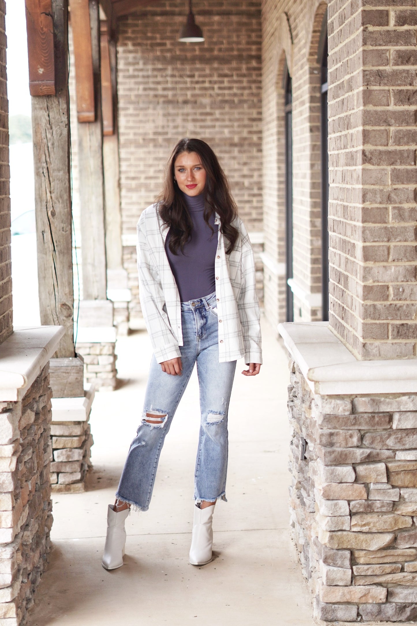 Harley High Rise Crop Flare Jeans High Rise Waistline Distressing on the Knee and the Hem of the Legs Light Denim Color Fitted Cropped Length 74% Cotton, 24% Polyester, 2% Spandex. Brunette model, white and grey flannel shirt, grey mockneck bodysuit, white booties. Stone and brick background.