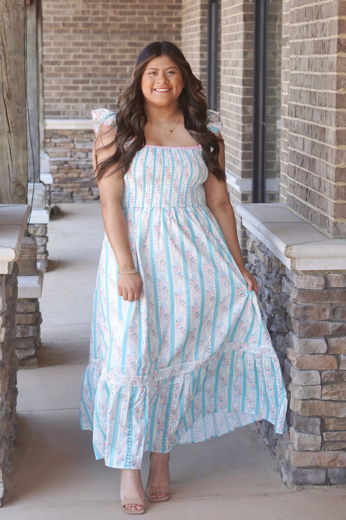 Woman wearing a pink and blue floral, sleeveless, maxi dress with ruffle sleeves, and it is smocked on the chest part of the dress. She has it paired with nude heels.