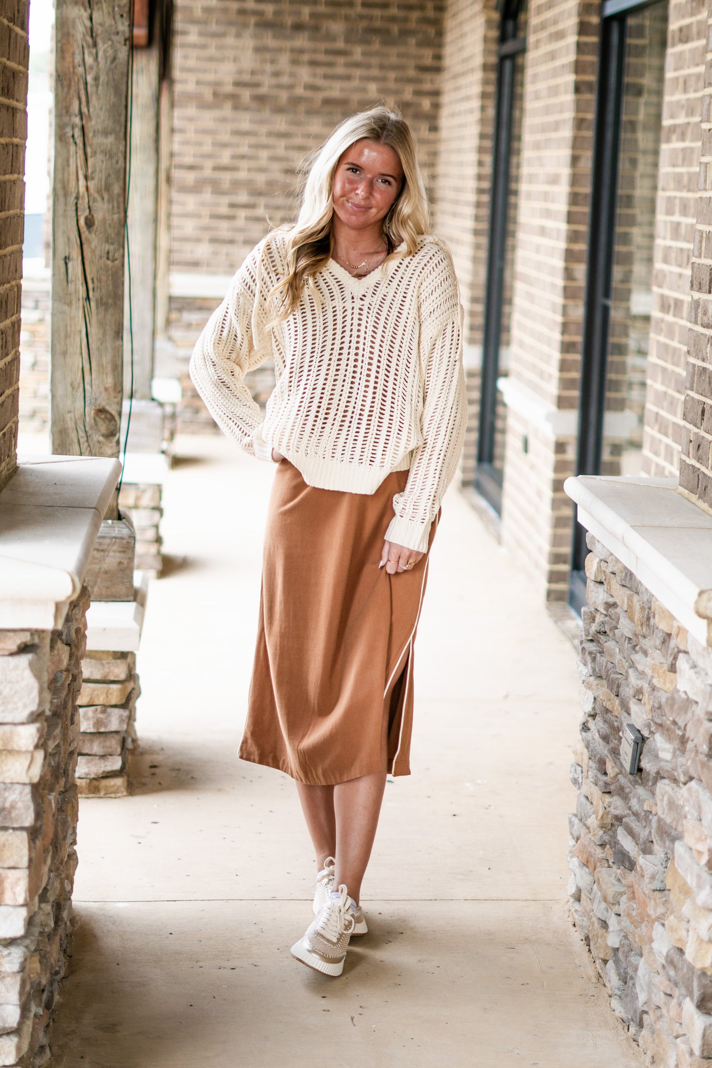 Olivia Open Knit Hooded Sweater Hooded Neckline Drawstrings Long Sleeves Open Knit Material Color/ Cream Waistline Length Relaxed Fit. Blonde model, tweed tennis shoes, sweater worn over burnt orange dress. Brick and stone background.