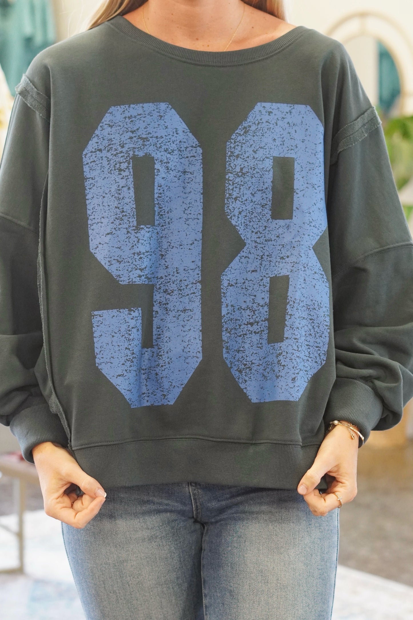 Crew neckline. High-low feature on the bottom of the hem. Dark grey color of the sweatshirt, "98" in blue. Long sleeves.