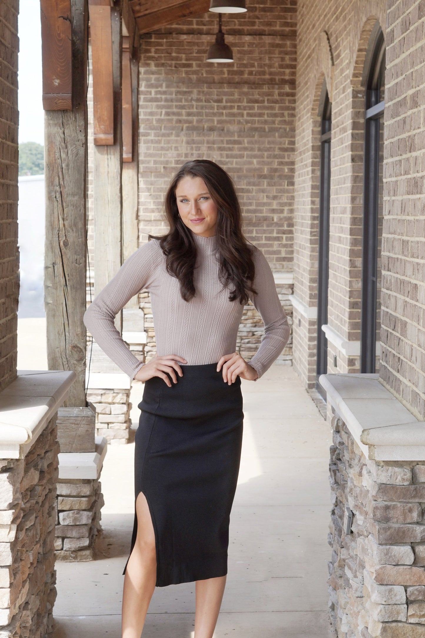 Pres Pointelle Mock Neck Sweater Mock Neckline Long Sleeves Pointelle Fabric Color: Tan Fitted Full Length. Brunette model, black skirt, stone and brick background.