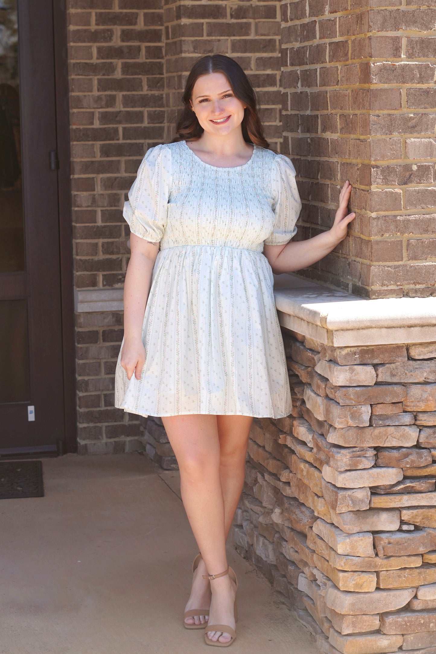 Woman wearing a floral, light blue, mini dress with puff sleeves. The top half of the dress is smocked and it flares out at the bottom. She has the dress paired with nude heels.