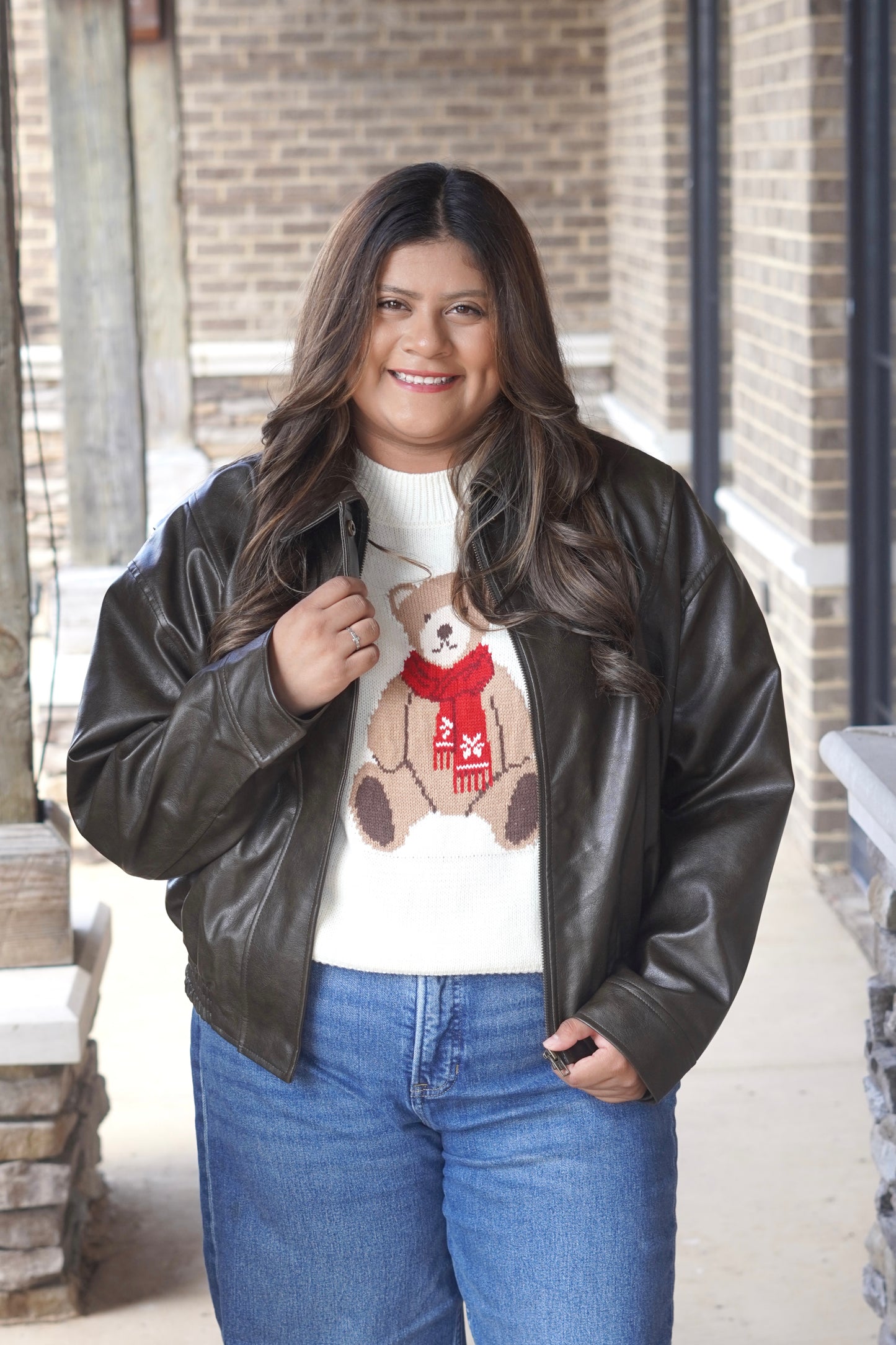 A woman wearing a dark brown faux-leather bomber jacket over a white knit sweater with a teddy bear graphic and blue jeans.