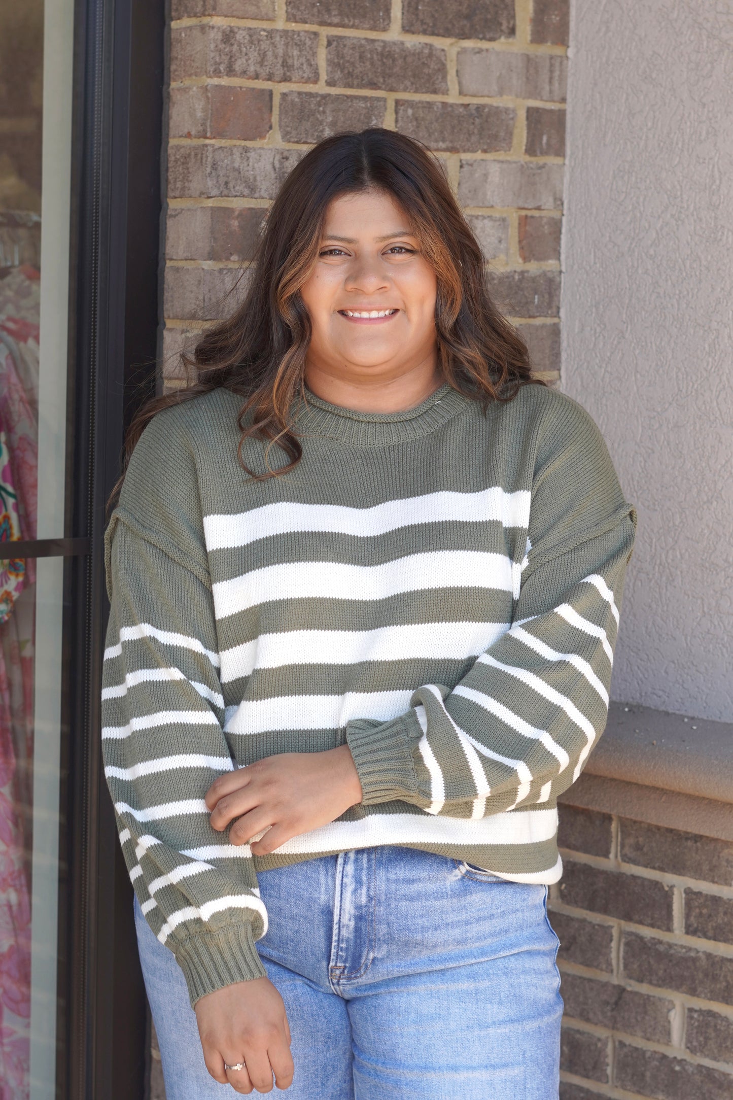 A woman wearing an olive green and white horizontally striped knit sweater with ribbed crew neck, cuffs, and hem, paired with light blue jeans.