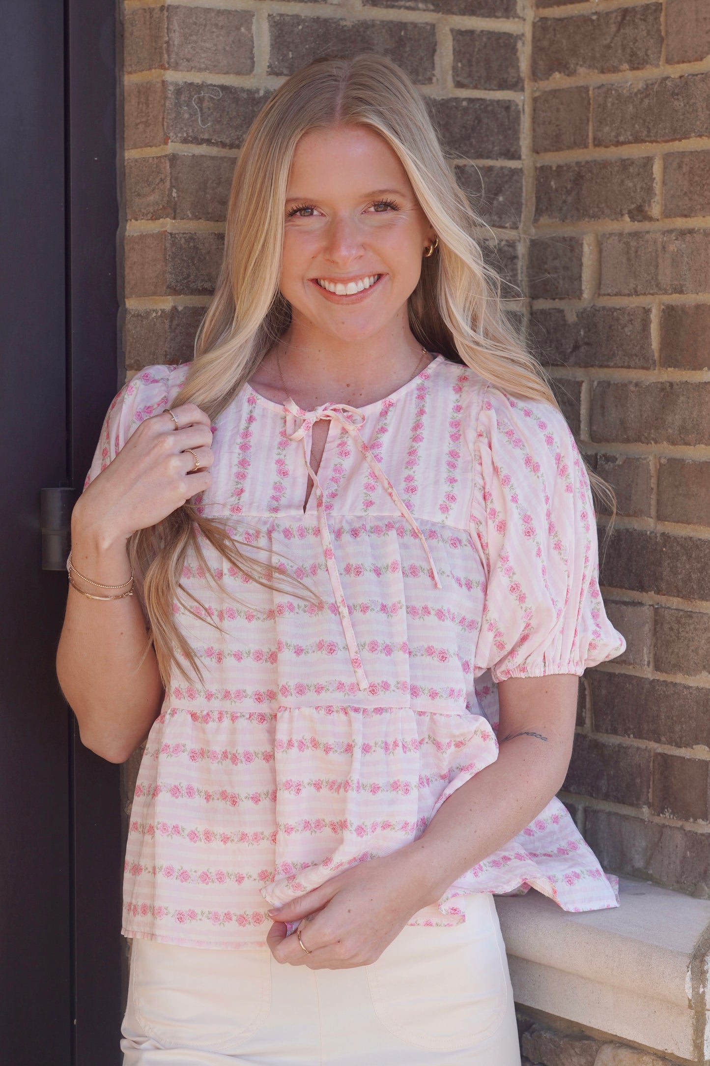 women wearing a pink floral striped print top, round neckline that ties into a bow, mid length puff sleeves, flowy.
