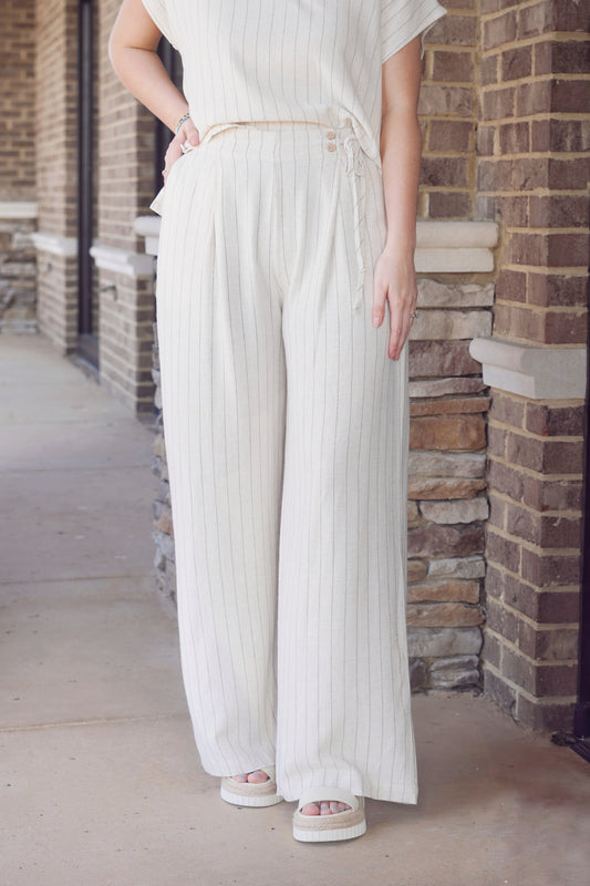 Women wearing a ivory flowy pants with thin black stripes and a elastic band that has buttons detailing on the side