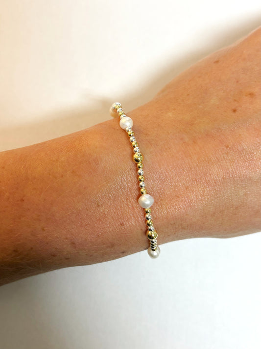 Pearl, sliver, gold bracelet on a wrist against a plain background