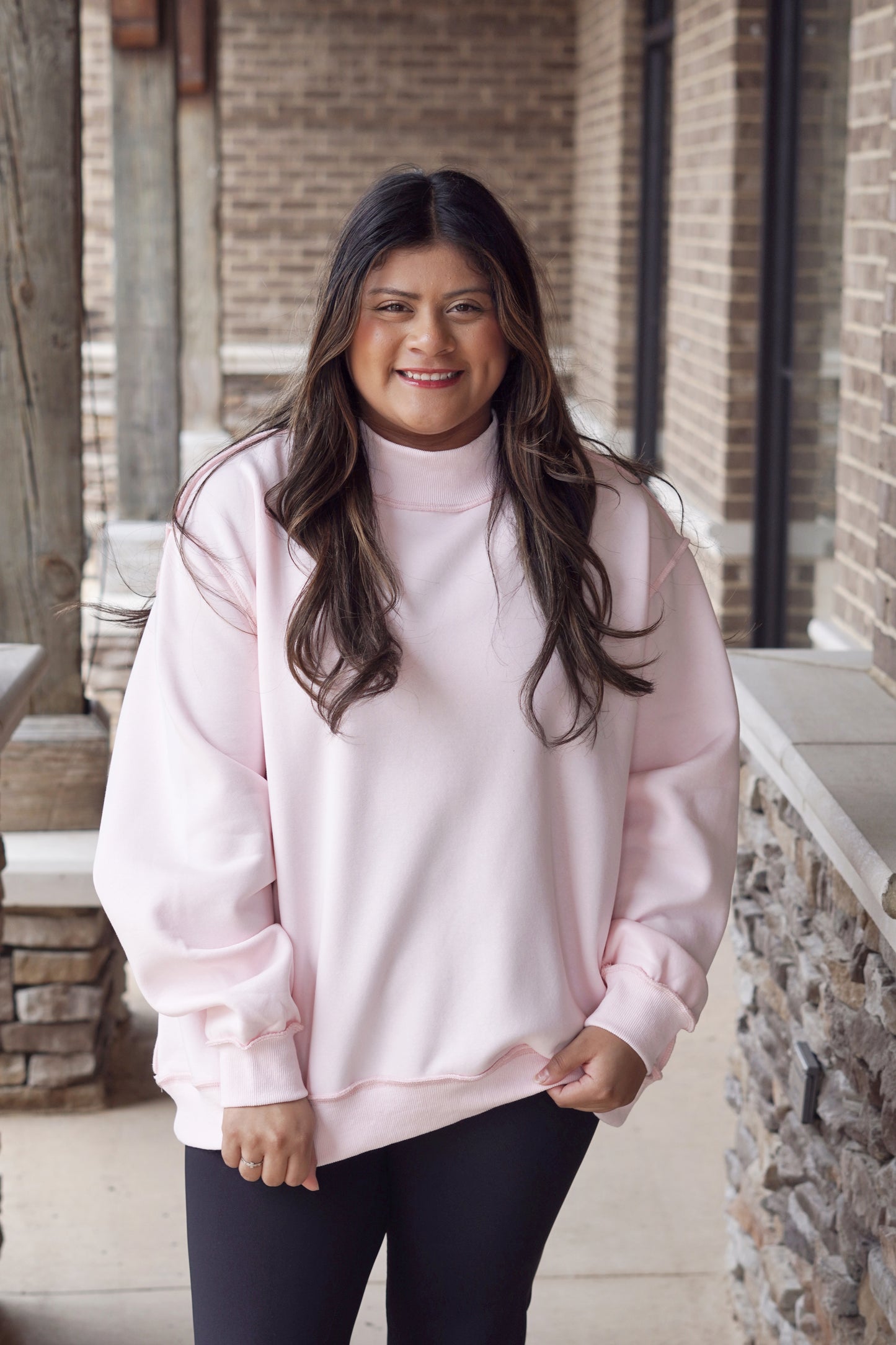 Woman wearing a baby pink oversized mock neck sweatshirt with black leggings.