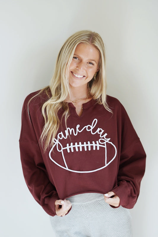 Gwen "Game Day" Cropped Crewneck Top Crew Neckline w/ Slit Long Cuffed Sleeves Raw Hem Cropped Length Maroon Color White Lettering Over a Football Relaxed Fit 60% Cotton, 40% Polyester