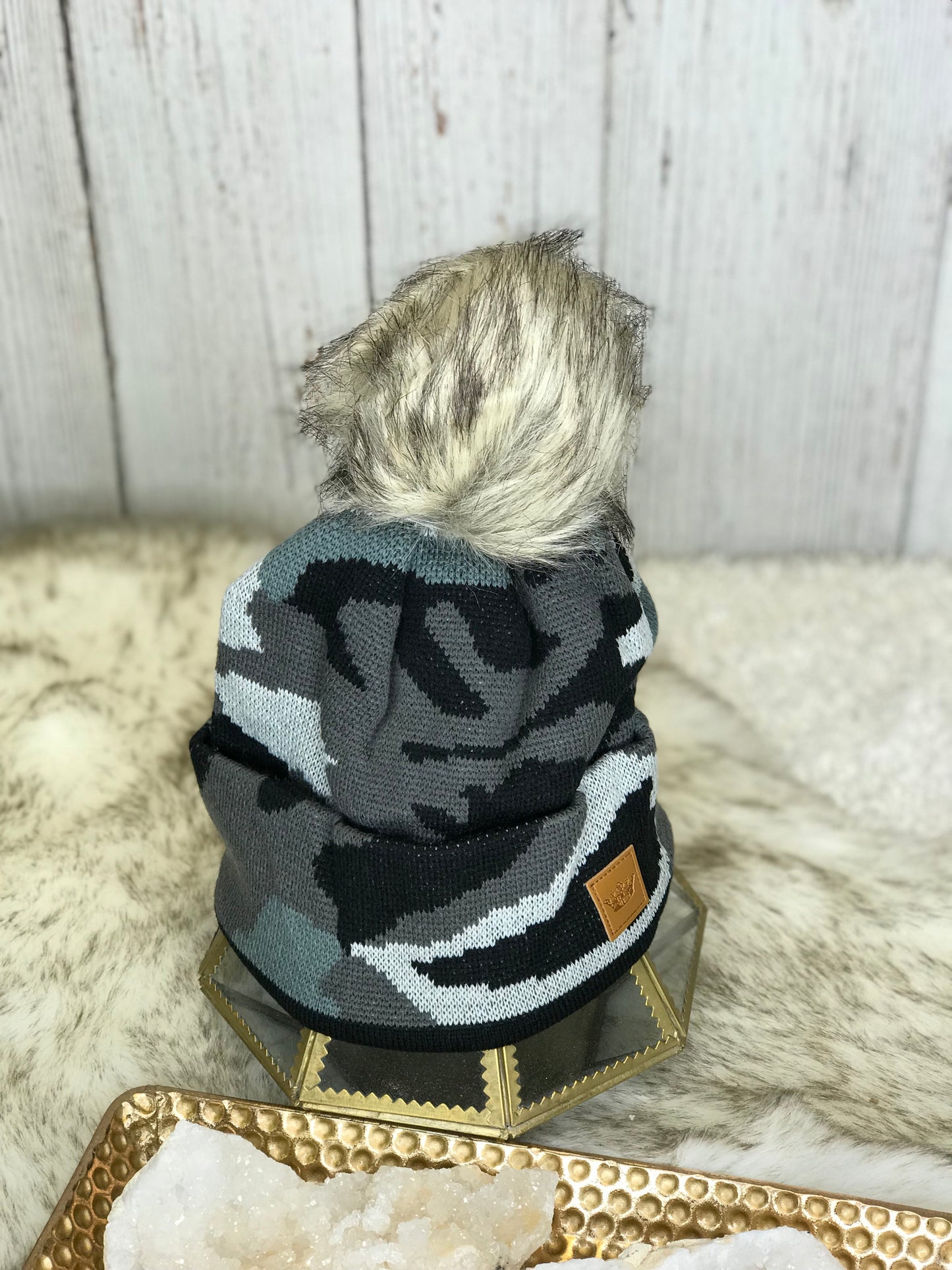 Camo toboggan with black and light and dark grey colors with a large cream and grey colored fur ball on the top of it