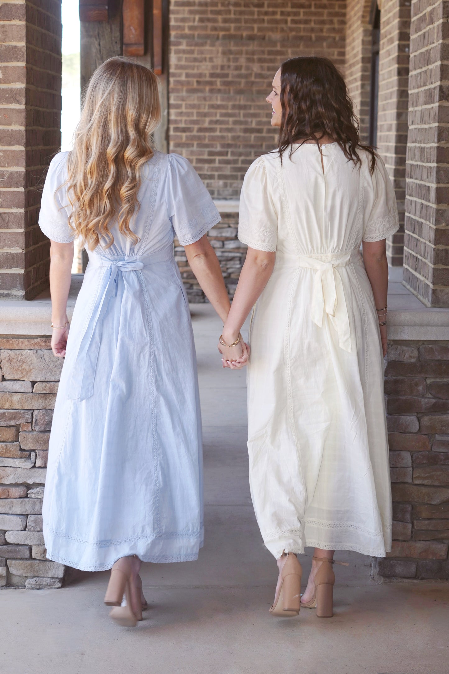 Two women wearing light blue and light yellow, short sleeve, maxi dresses, that have belts that tie in the back.