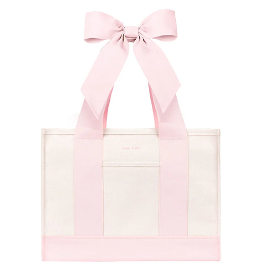 A light pink and white tote bag featuring wide pink shoulder straps tied into a large bow at the top, with a front pocket and a solid pink base trim.
