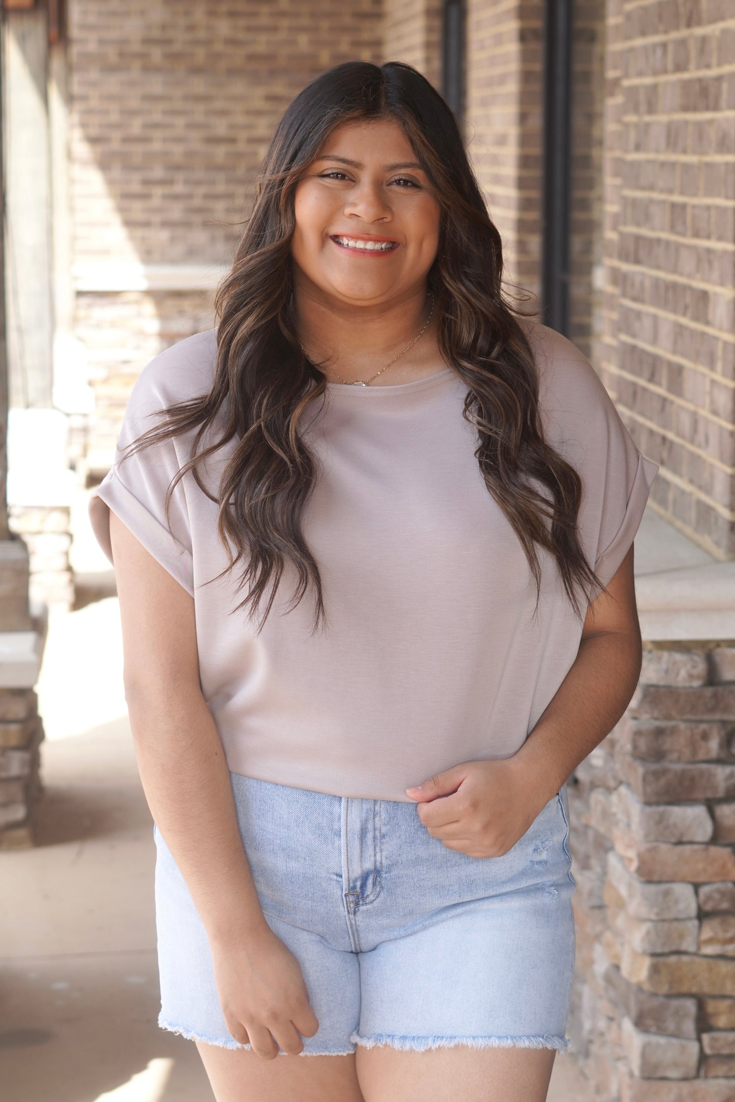 Woman wearing a tan colored short cap sleeve top with a round neckline paired with blue jean shorts