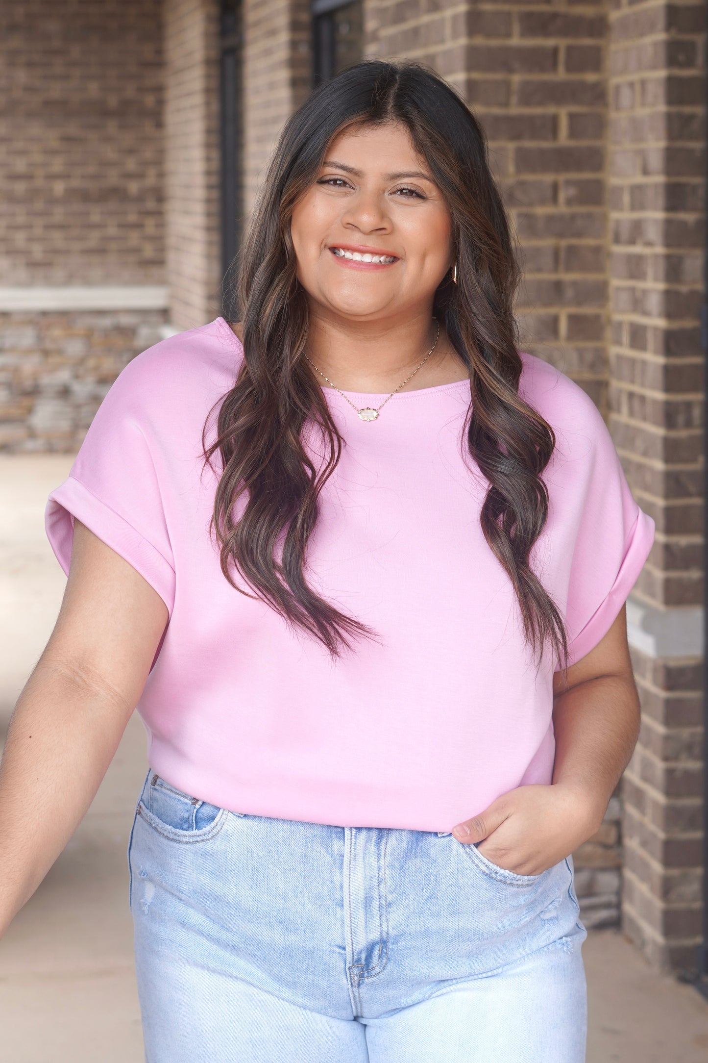Woman wearing a short sleeve pink top with a round neckline and rolled sleeves paired with blue jeans