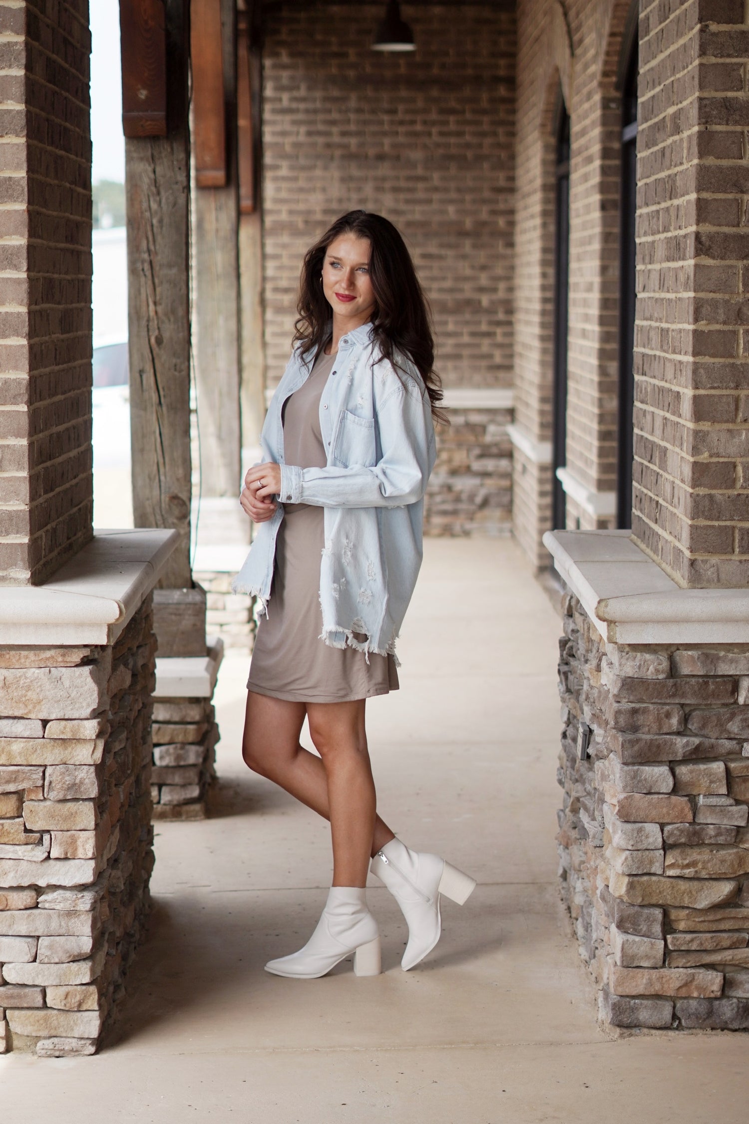 Darla Denim Distressed Shirt Collared Neckline Long Cuffed Sleeves Distressing Light Denim Color Front Left Pocket Fringed Hem Line Relaxed Fit Upper Thigh Length 100% Cotton. Brunette model, nude dress, white booties. Stone and brick background.