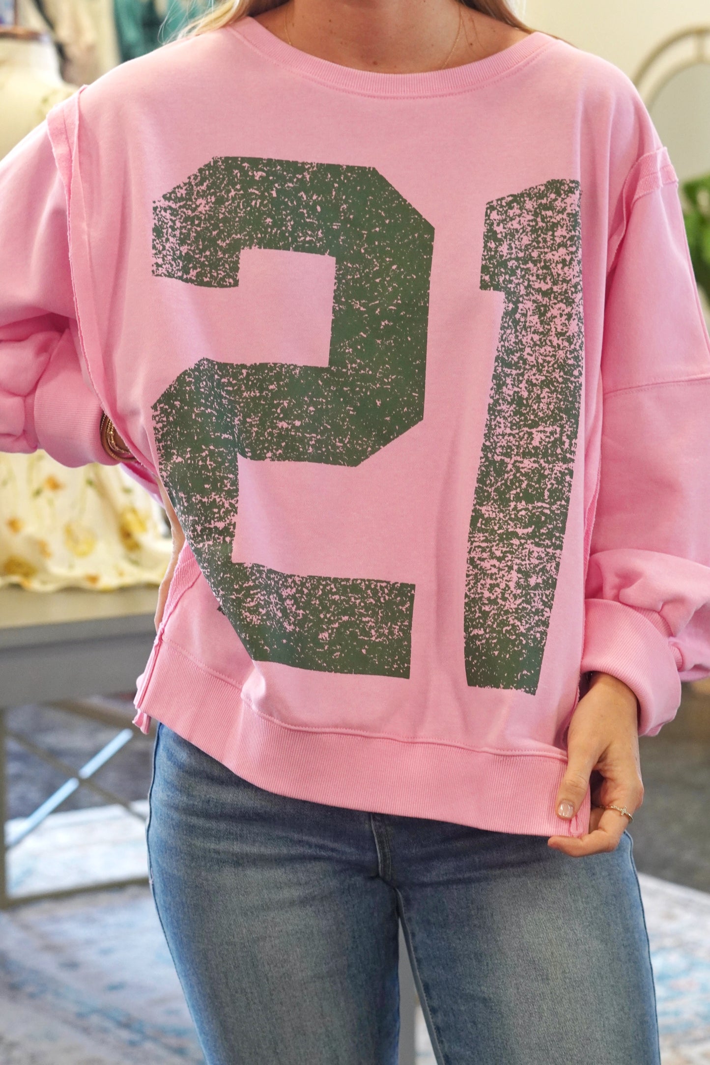 Crew neckline. High-low feature on the bottom of the hem. Light pink color of the sweatshirt, "21" in dark green. Long sleeves. 