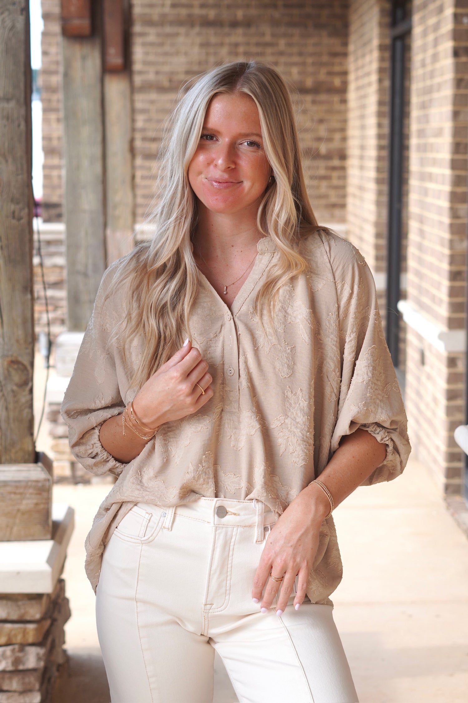 Women wearing a Taupe Short Sleeve Flowy Blouse, V-Neckline, Half Button Down, Full Length, textured print, paired with some ivory color jeans. 