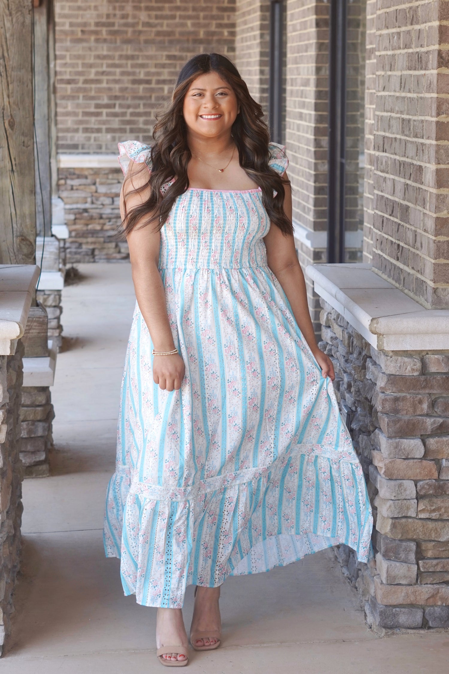 Woman wearing a pink and blue floral, sleeveless, maxi dress with ruffle sleeves, and it is smocked on the chest part of the dress. She has it paired with nude heels.