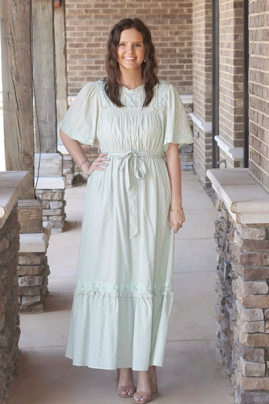Woman wearing a light green, maxi dress with lace detailing and belt that ties around the waist, paired with nude heels.