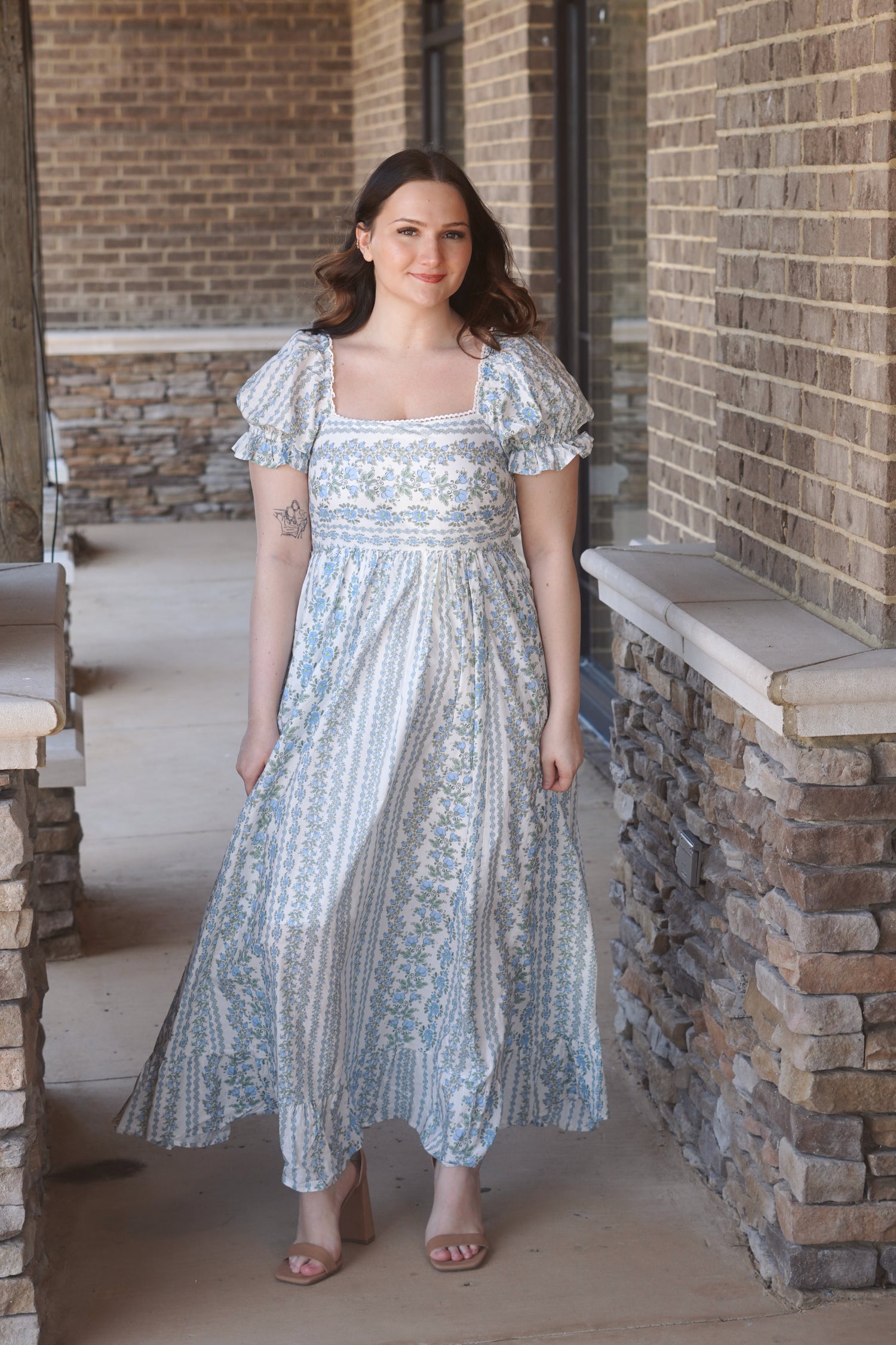 A women wearing a blue and white maxi dress with a floral pattern, puff sleeves, and a square neckline.
