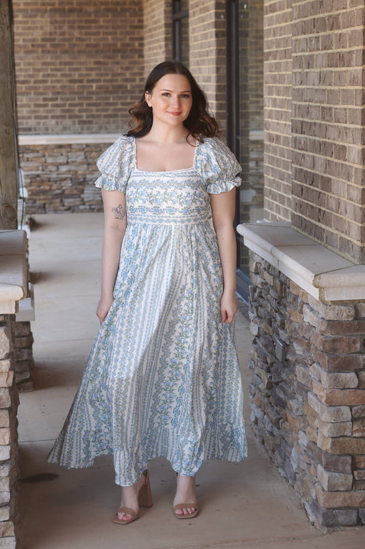 A women wearing a blue and white maxi dress with a floral pattern, puff sleeves, and a square neckline.