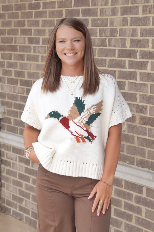 A woman wearing a cream knit short-sleeve sweater with a colorful duck on the front, paired with brown pants.