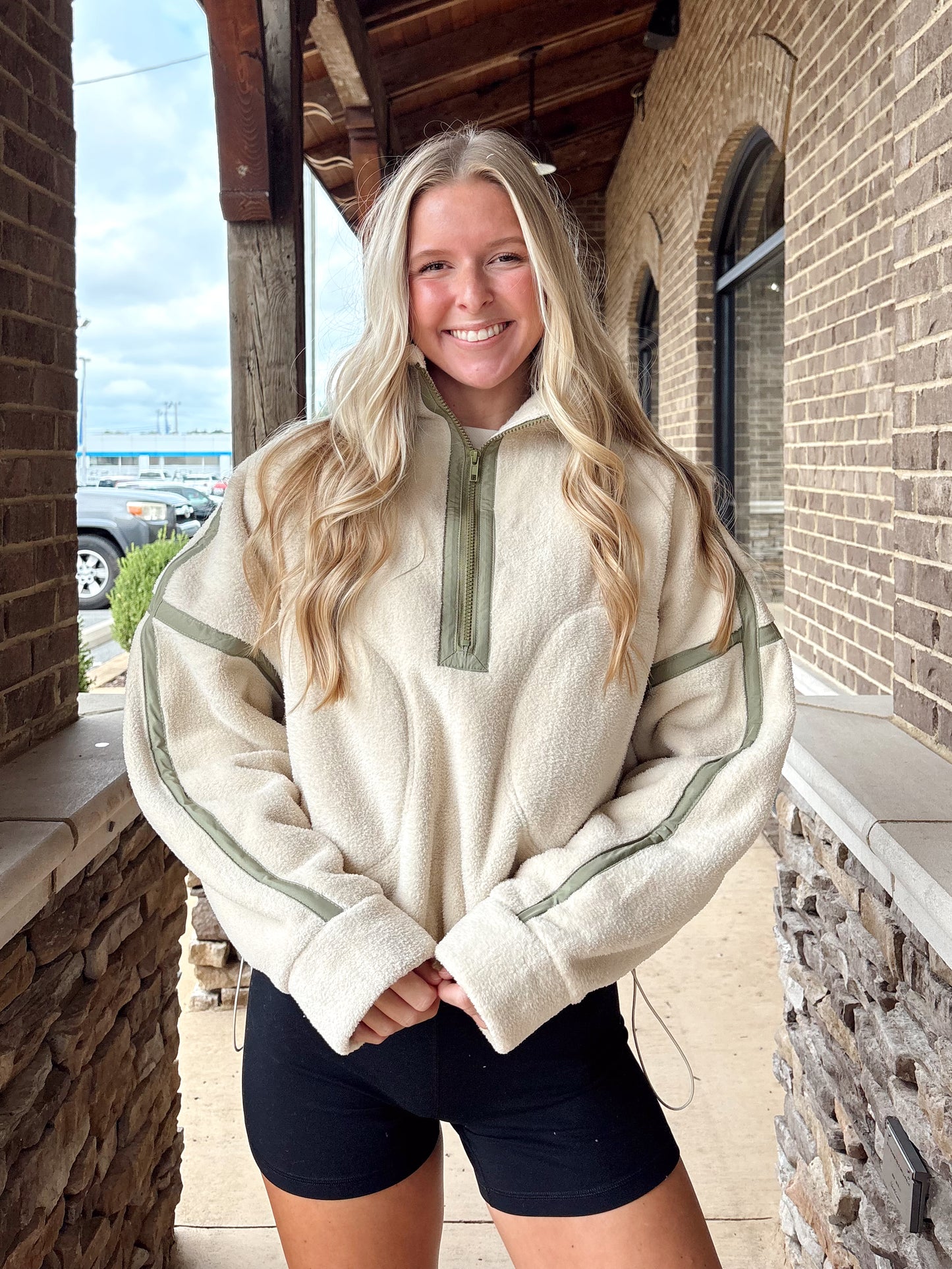 Woman wearing a cream-colored fleece quarter-zip pullover with sage green stripe details on the sleeves, paired with black athletic shorts.