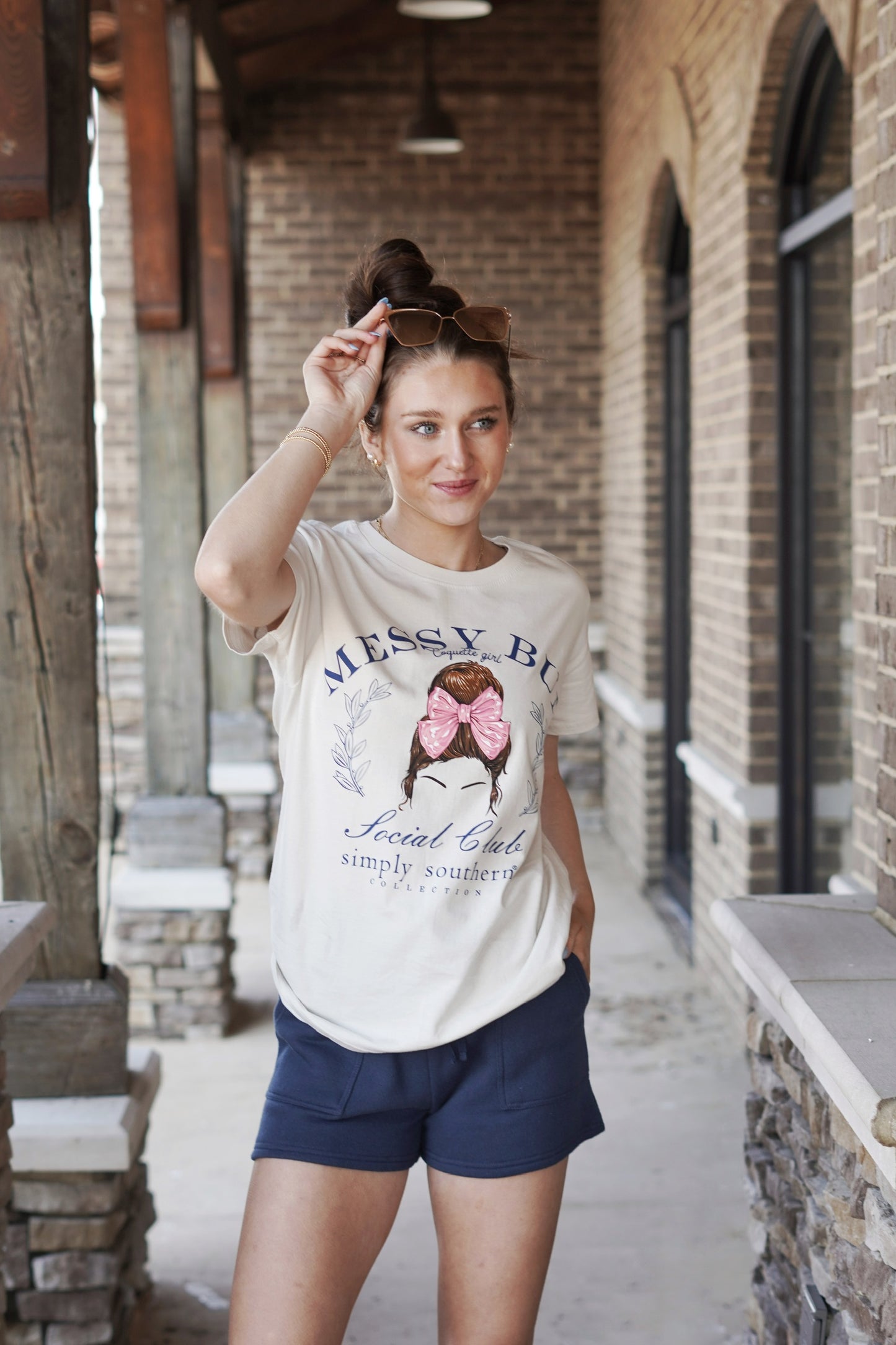 Wisp tan short sleeve shirt with the phrases 'Messy Bun', Coquette Girl', and 'Social Club'. There is a messy bun hairstyle with a pink bow. Simply Southern logo below and on upper back<ul> <li>Crew Neckline</li> <li>Short Sleeves</li> <li>Full Length</li> <li>Color: Cream</li> <li>100% Cotton</li> </ul>