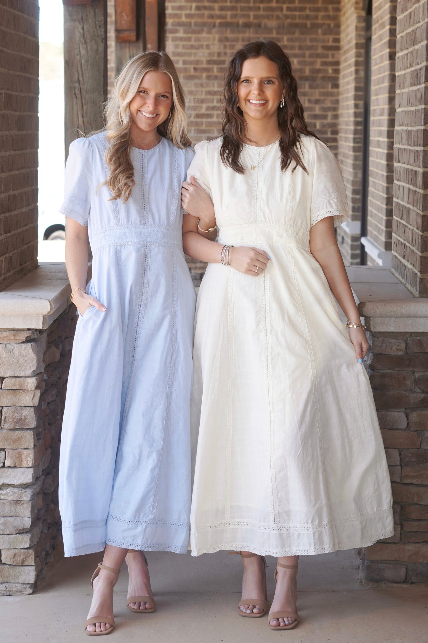 Two women wearing light blue and light yellow, short sleeve, maxi dresses, that have lace detailing and a round high neckline, paired with nude heels.