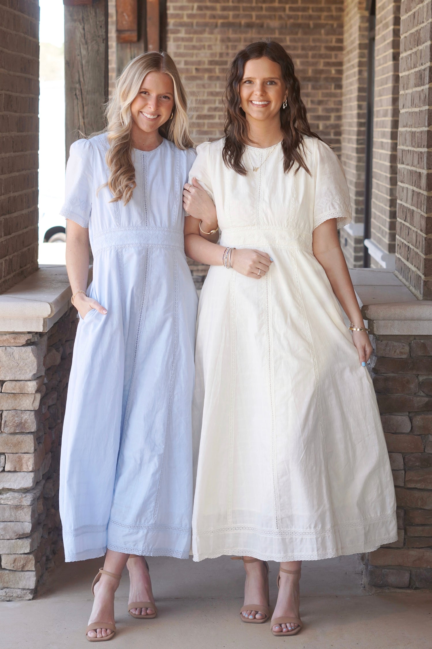 Two women wearing light blue and light yellow, short sleeve, maxi dresses, that have lace detailing and a round high neckline, paired with nude heels.