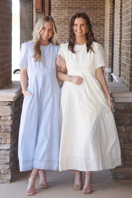 Two women wearing light blue and light yellow, short sleeve, maxi dresses, that have lace detailing and a round high neckline, paired with nude heels.