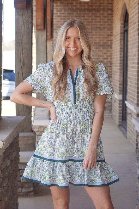 Woman wearing a short-sleeved, floral-patterned dress. The dress has a white base with a blue and green floral print. It has a elastic fitted waist and a ruffled hem, with green/ blue trim along the v neckline and hem, it is a mid thigh length dress.