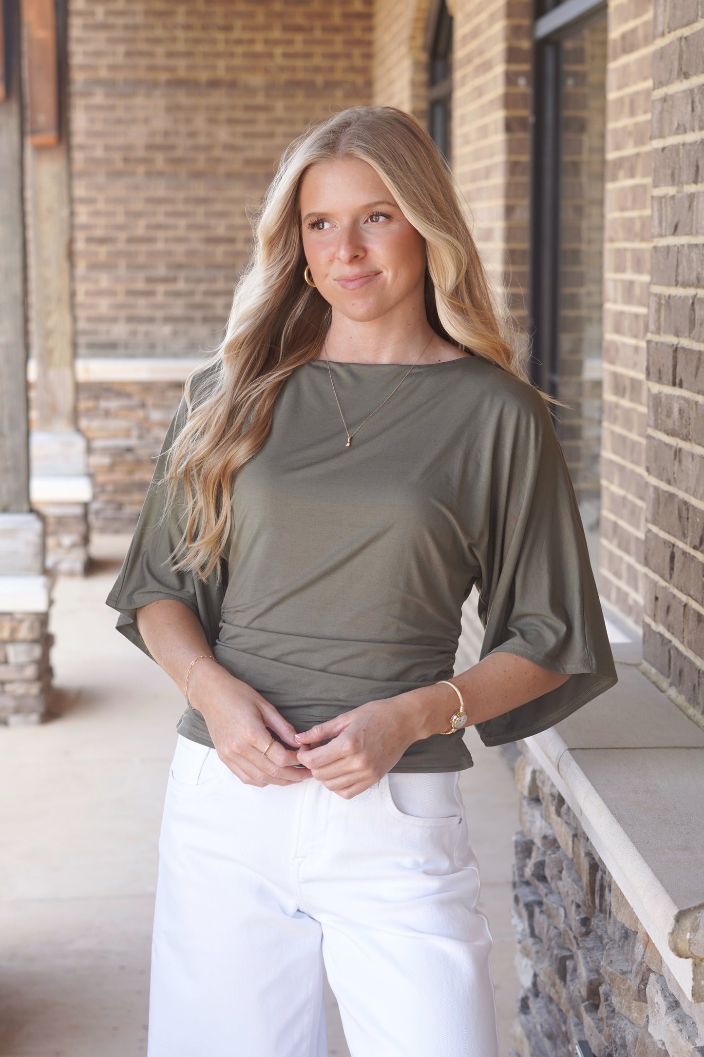 Woman wearing a green top, with cinch waist and mid-length flowy  sleeves and white pants.