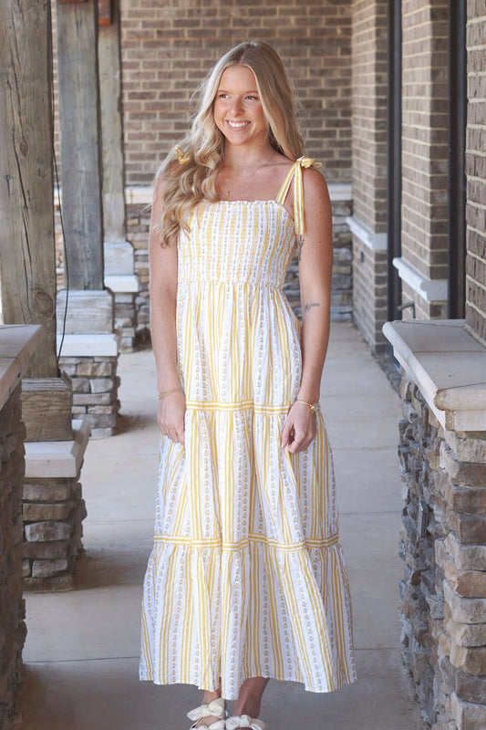 Woman wearing a yellow, striped, tiered maxi dress with a floral pattern and straps that tie.