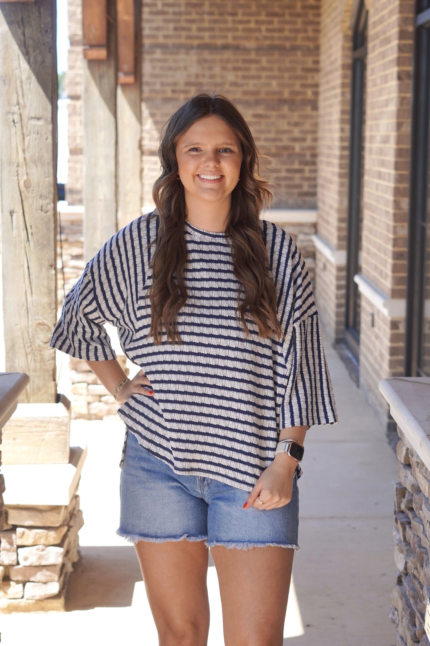 Women wearing a navy Textured Striped Top, 3/4 Length Sleeve, Relaxed Fit paired with blue jean shorts. 

