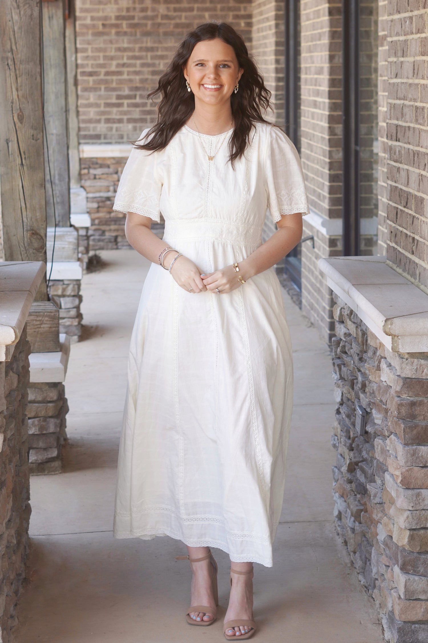 Women wearing a light yellow, short sleeve, maxi dress, that have lace detailing and a round high neckline, paired with nude heels.