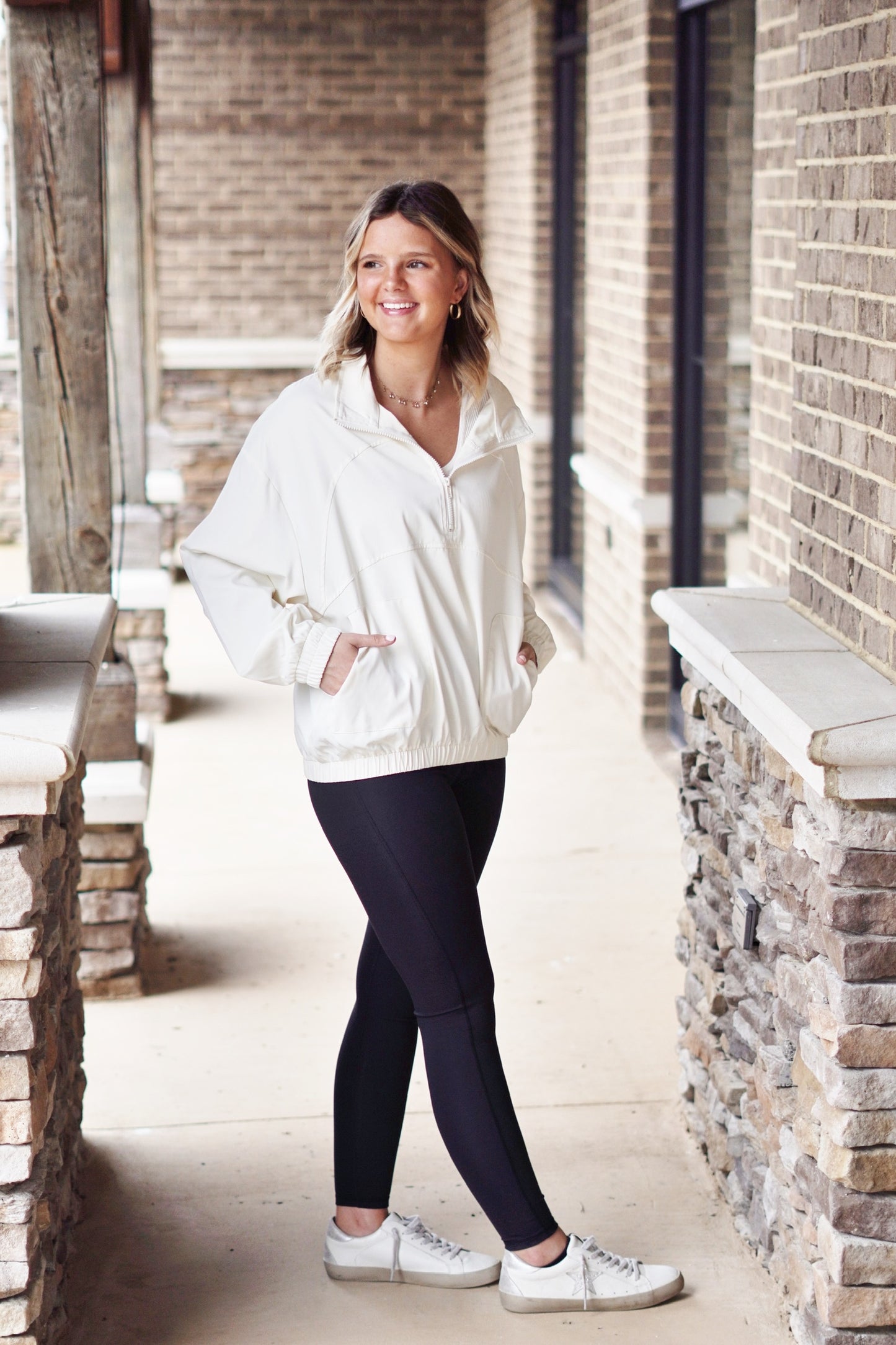 Hannah Half Zip Pullover Half Zip, Collar neckline Color/ Soft Beige Long Sleeve Elastic Band On End Of Sleeves And Waistline Two Front Pockets Relaxed Fit Full Length 100% Polyester. Blonde model, black leggings, sneakers. Brick and stone background.