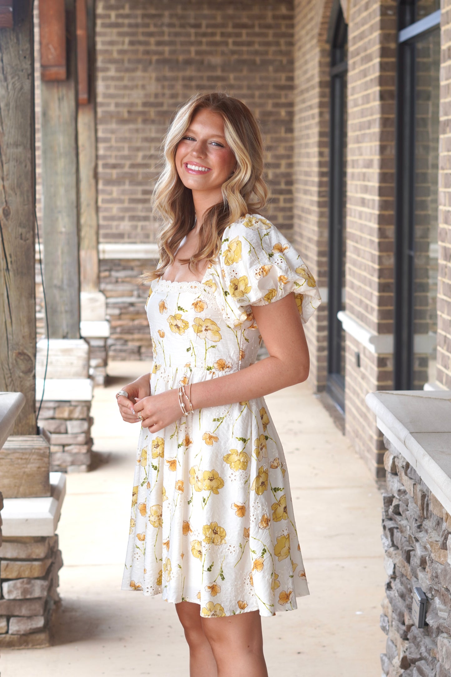 Women wearing an ivory dress with yellow flowers print, above the knee length, short puff sleeves, square neckline. 
