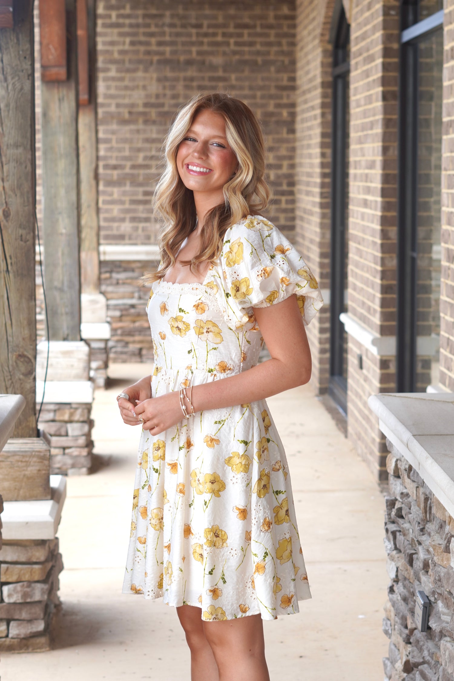 Women wearing an ivory dress with yellow flowers print, above the knee length, short puff sleeves, square neckline. 