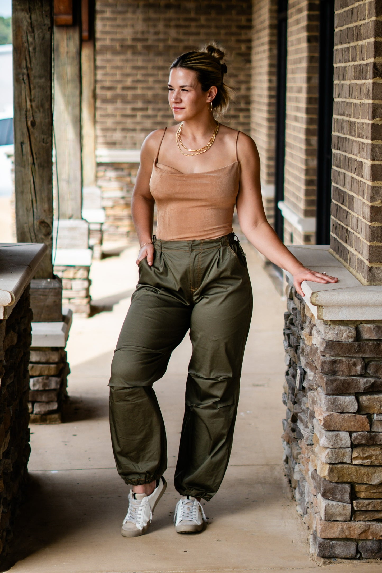 Polly Parachute Jogger Pants Elastic Waistband w/ Zipper Fly and Button Closure Full Length Legs Cinched at the Ankle Pockets Dark Olive Color. Blonde model, tan bodysuit, sneakers. Stone and brick background.