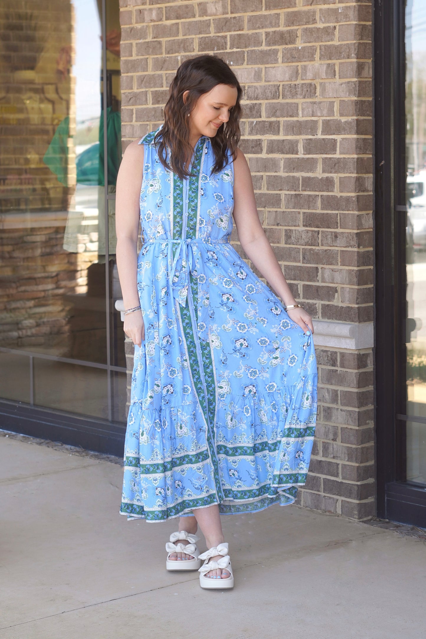 Woman wearing a sleeveless, collared, button up, blue and green maxi dress with a belt that ties paired with cream sandals.