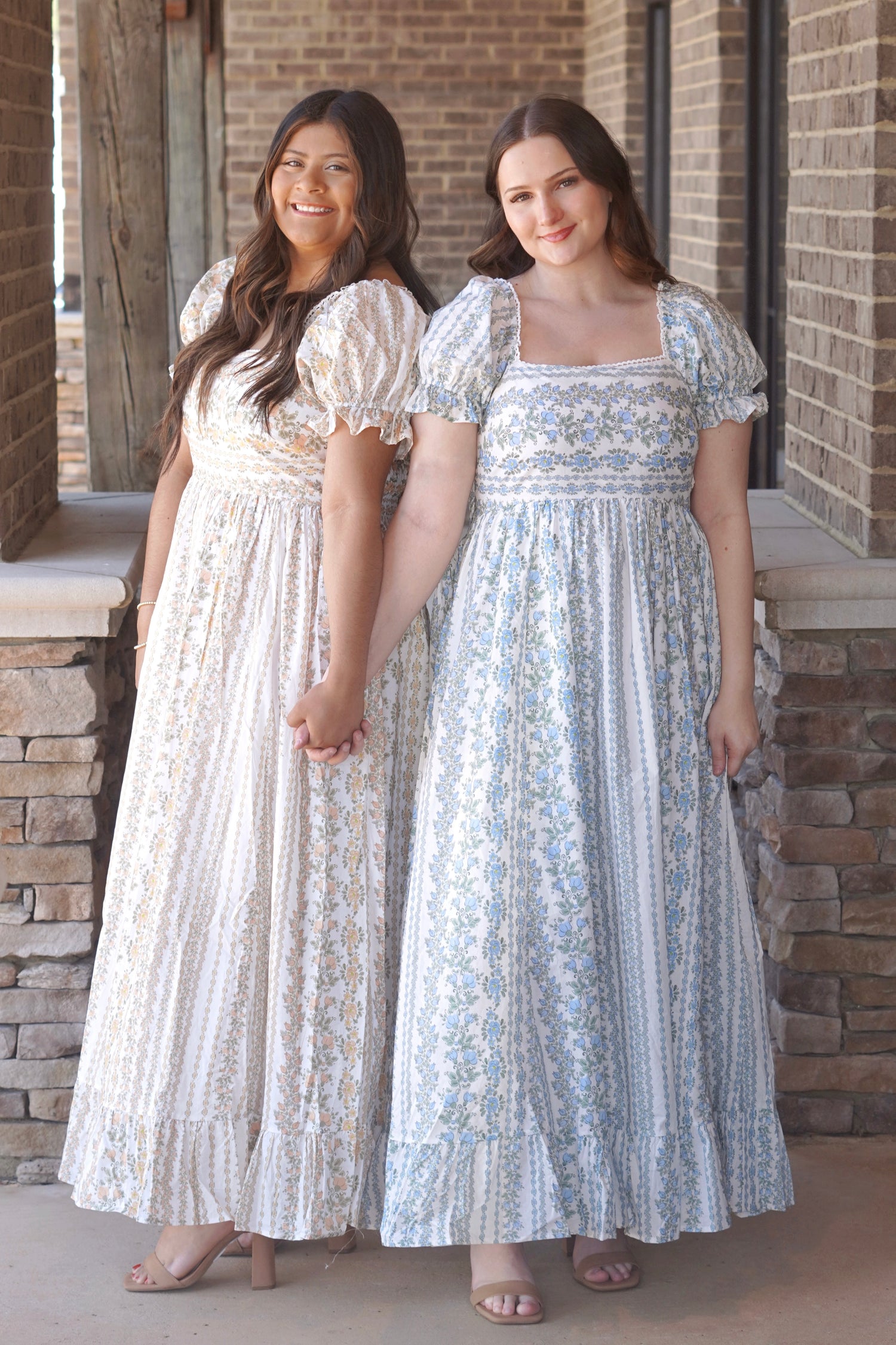 Two women wearing blue and yellow maxi dresses with a floral pattern, puff sleeves, and a square neckline.