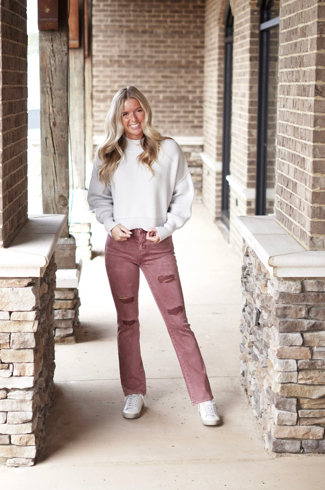 Darling Dusty Rose Cropped Straight Leg Jeans Zipper Fly Straight Leg Cropped Length Fitted Minor Distressing Dusty Rose Color 93% Cotton, 5% Polyester, 2% Spandex. Blonde model, grey sweater, sneakers. Stone and brick background.