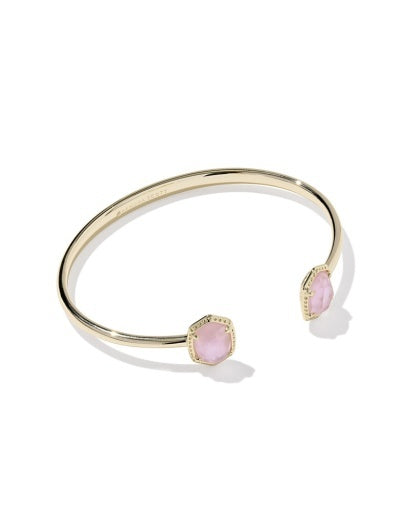 Gold open cuff bracelet with pink stone at the end in a gold texture frame. 