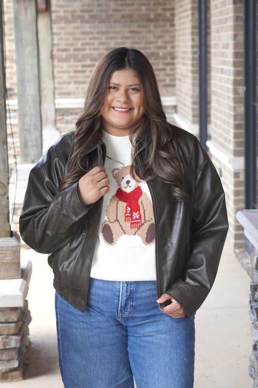 A woman wearing a dark brown faux-leather bomber jacket over a white knit sweater with a teddy bear graphic and blue jeans.
