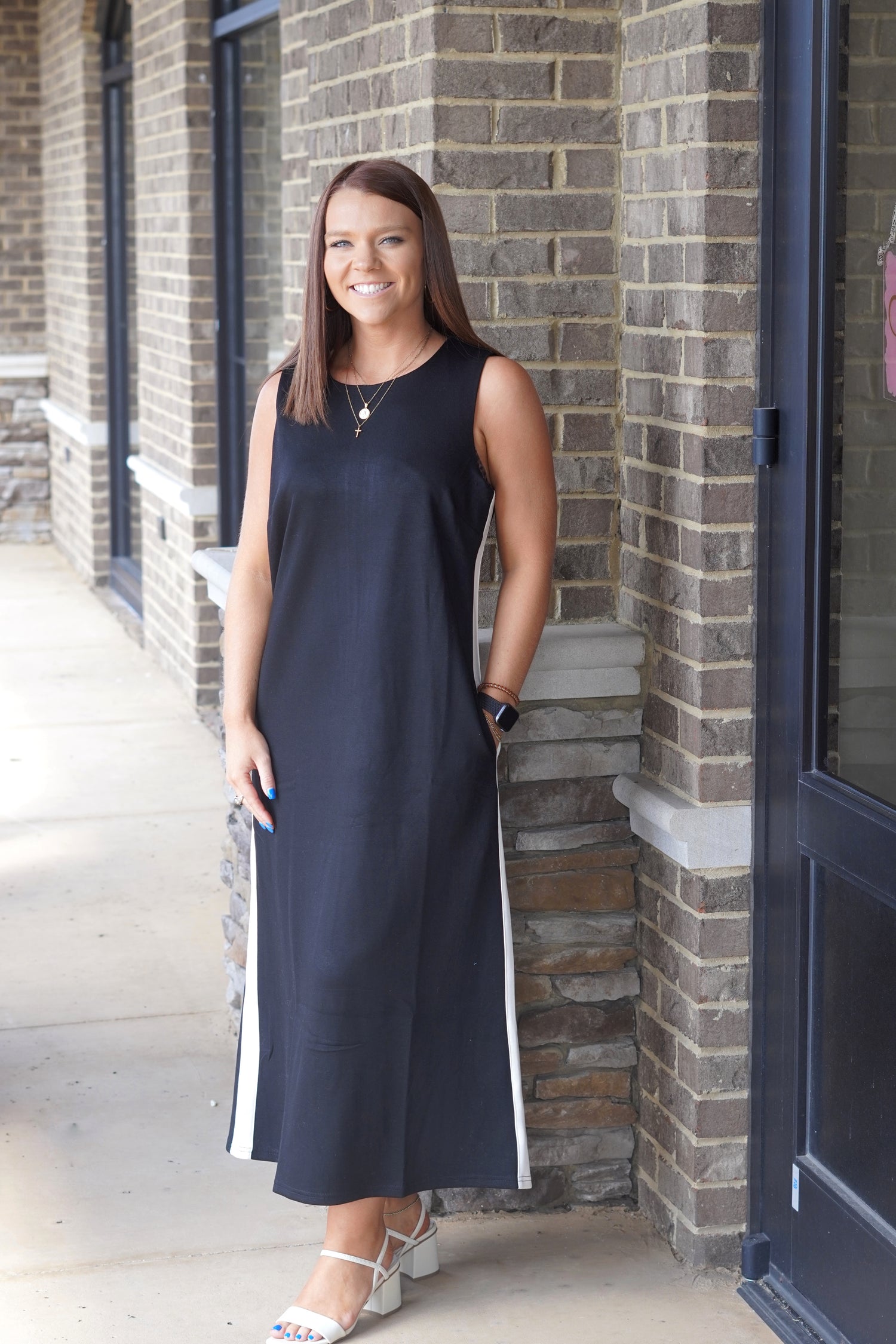 A woman wearing a sleeveless black midi dress with white side stripes, standing outdoors against a brick building wall.