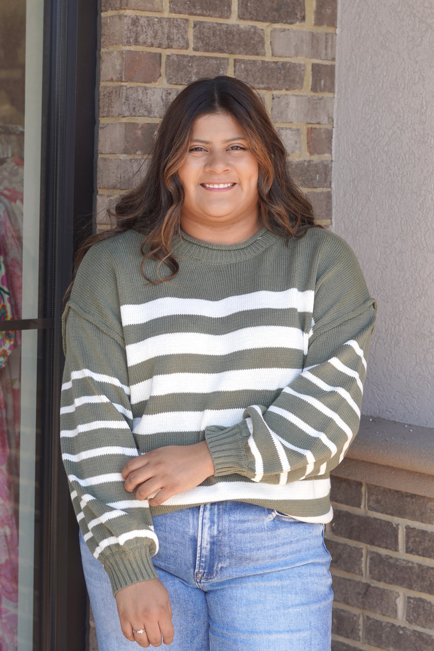 A woman wearing an olive green and white horizontally striped knit sweater with ribbed crew neck, cuffs, and hem, paired with light blue jeans.