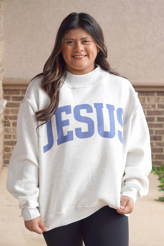 Woman wearing a ash grey oversized mock neck sweatshirt that has "Jesus" written in blue with black leggings. 